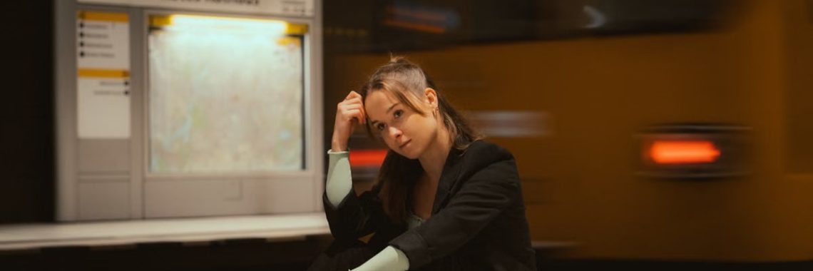 A young woman in casual attire seated at a Berlin subway station with trains in motion.